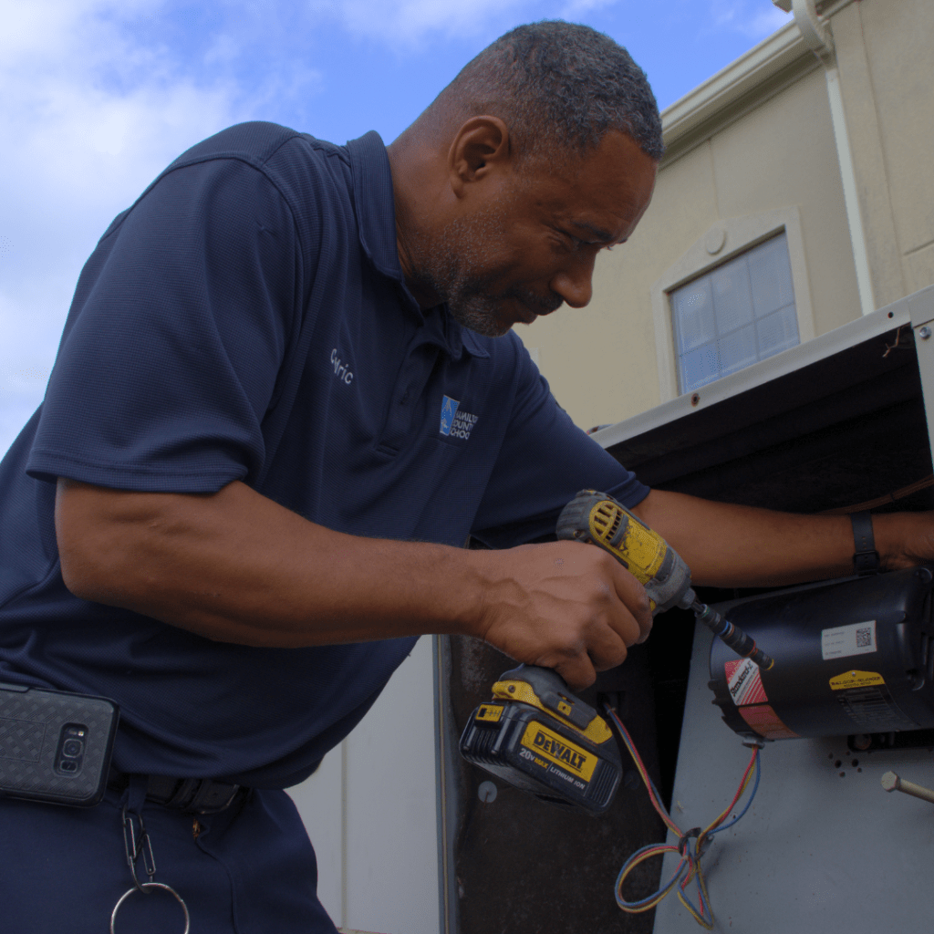 Maintenance Worker Outside A maintenance worker adjusting an air conditioning unit at a HCS school, ensuring the system is running properly to keep classrooms comfortable